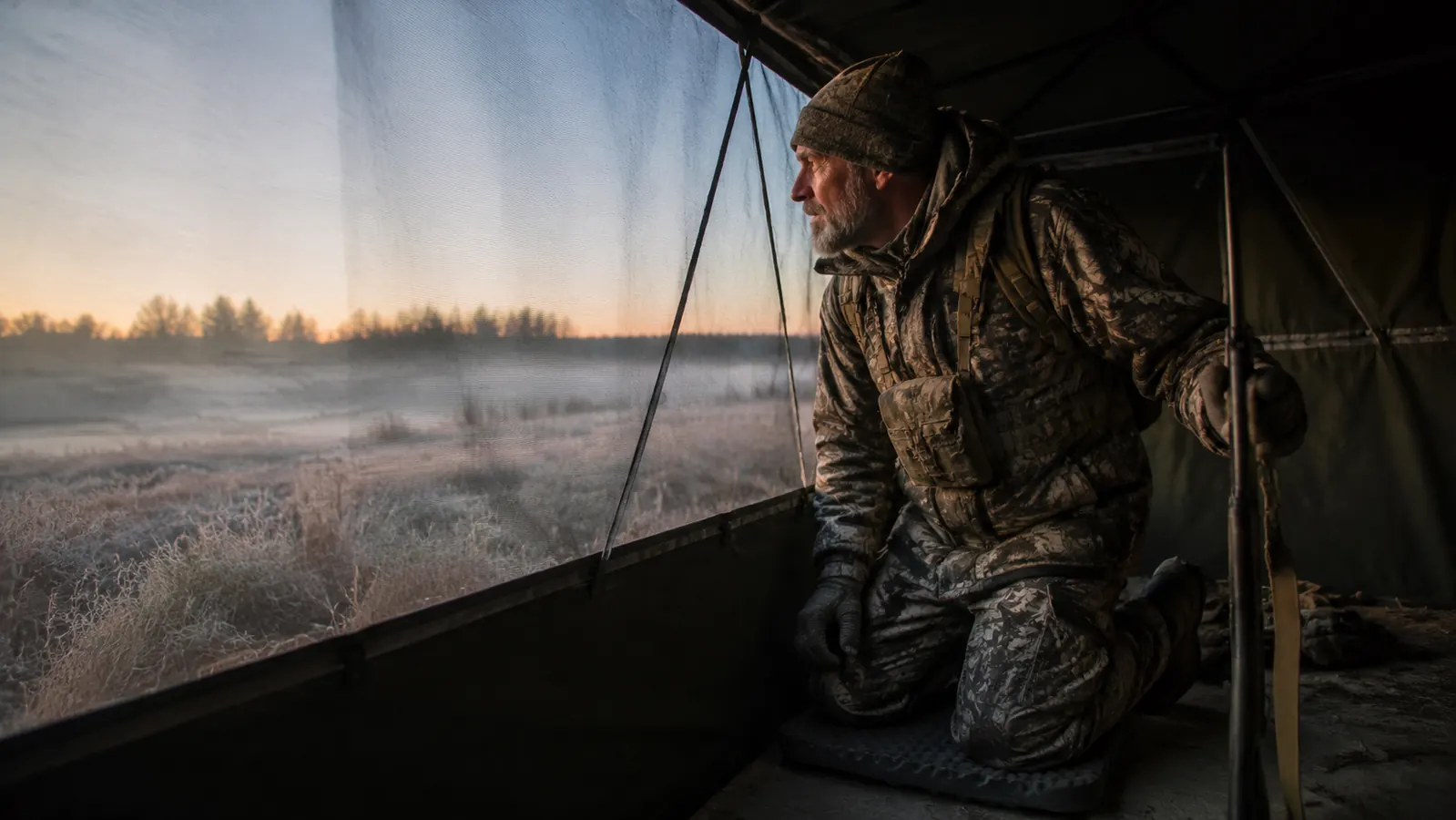 An older hunter kneeling on a foam pad inside a ground blind at dawn, looking out over a frosty field
